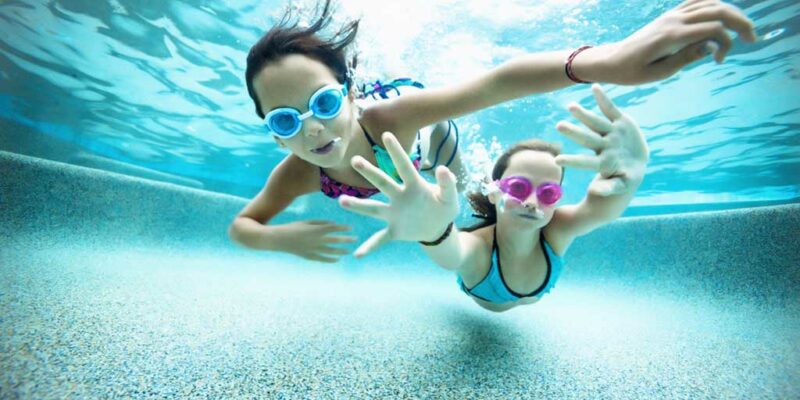 two girls swimming in a pool together underwater each wearing a pair of goggles