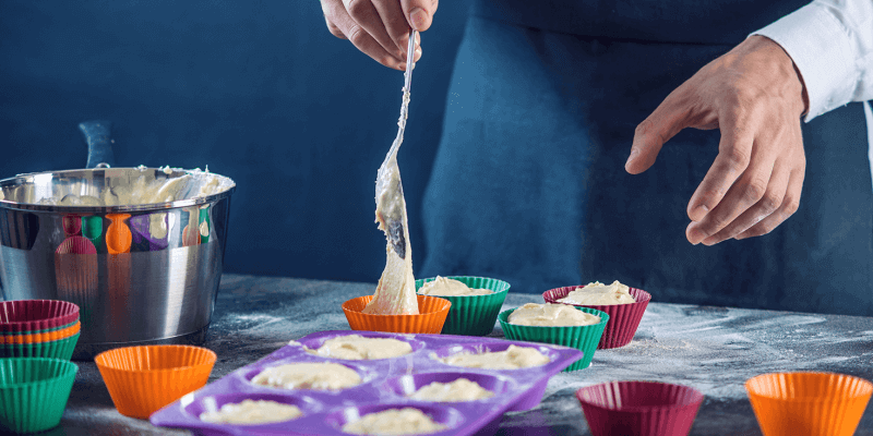 Chef pastry in a black apron filling the dough into a cupcake silicone mold on dark background.